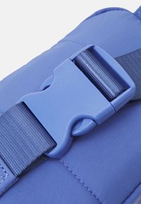 Gros plan sur une boucle à déblocage latéral bleue en plastique fixant une sangle tissée sur une surface en tissu bleu rembourré.