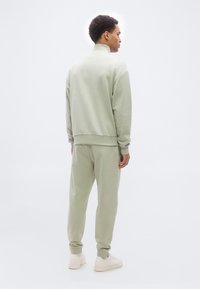 Άνδρας που στέκεται γυρισμένος από μπροστά, φοράει ανοιχτοπράσινο φούτερ με ψηλό λαιμό και ταιριαστά φόρμα παντελόνια, με λευκά αθλητικά παπούτσια, σε απλό λευκό φόντο.