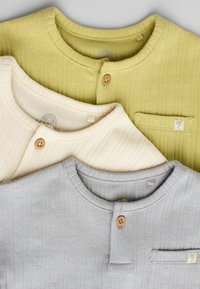 Trois chemises côtelées en vert olive, crème et gris clair. Chacune présente un col rond, des boutons en bois et un petit détail de poche.