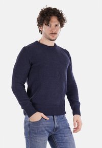 Maglione lavorato a maglia blu navy con collo a giro e polsini a coste. Indossato sopra jeans azzurri chiari, con una vestibilità comoda.