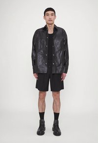 Juoda odinė marškinių striukė su spaudėmis, dėvima ant juodos aptemtos marškinėlės ir juodų šortų. Modelis taip pat dėvi juodus aulinukus ir kojines.