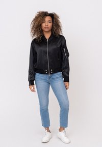 Zwarte satin bomberjack met een ritssluiting aan de voorkant, geribde manchetten en zoom; gecombineerd met lichtblauwe enkellange jeans en witte sneakers.