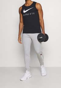 Top sin mangas negro de Nike con el texto "ATLETA", pantalones de chándal ajustados gris y zapatillas deportivas blancas, sosteniendo una mancuerna negra en una mano.