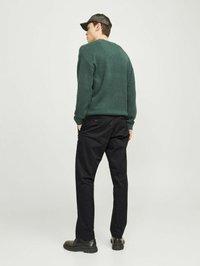 Grønt sweater med en struktureret finish, sorte bukser og sorte sko. Modellen har en kasket på og er placeret med en let drejning.