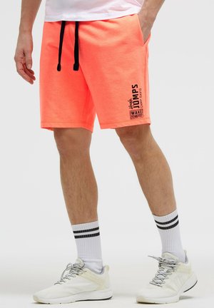 Mann trägt leuchtend orangefarbene Shorts mit schwarzem Kordelzug, weißen Socken mit schwarzen Streifen und weißen Sportschuhen, steht mit einer Hand in der Tasche.