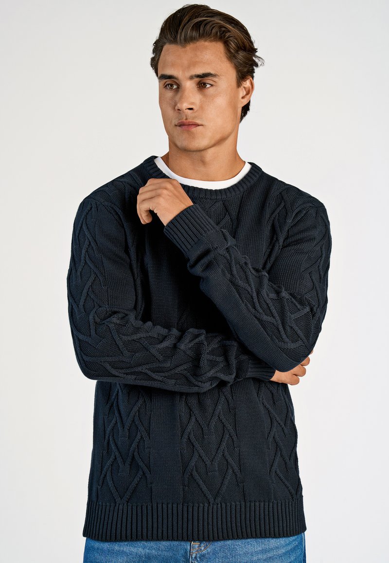 Dunkelblauer Strickpullover mit Zopfmuster, gerippten Bündchen und Saum, über einem weißen Hemd getragen, in lockerem Schnitt.