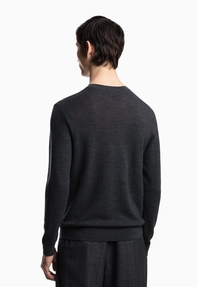 Uomo con capelli corti scuri che indossa un maglione grigio scuro a trama e pantaloni grigio scuro, visto di spalle su uno sfondo bianco.