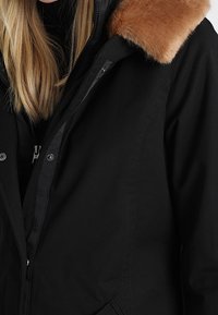 Czarna ocieplana kurtka z kapturem wyłożonym futrem, zapinana na zamek, z kieszeniami po bokach. Tkanina ma gładką teksturę i dopasowany krój.