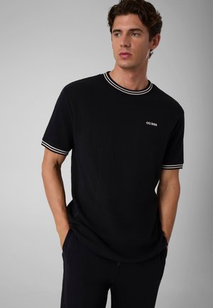 Junger Mann mit braunen Haaren trägt ein schwarzes Guess-T-Shirt mit weiß gestreiftem Kragen und Ärmelbündchen, Hände in den Taschen, schaut nach rechts.