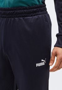 Άτομο που φοράει μαύρο παντελόνι φόρμας Puma με ελαστική μέση και εμφανές λογότυπο της μάρκας κοντά στην τσέπη, με το χέρι χαλαρό στο πλάι.