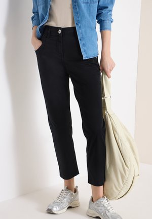 Persona che indossa pantaloni neri corti, sneakers argento, camicia di denim blu e tiene una grande borsa beige di fianco.