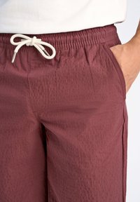 Burgundy dragsko byxor i texturerat tyg. Har en elastisk midjeband och sidofickor. Kontrastvit dragsko med metalländar.