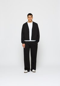 Fekete, rövid fazonú kardigán gombokkal és raglánujjakkal, laza fekete nadrággal párosítva. Fehér, kerek nyakú póló látszik alatta.