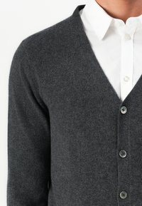 Cardigan in lana grigio scuro con bottoni indossato sopra una camicia bianca con colletto, mostrato dalla gola fino a metà busto.