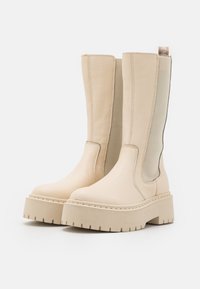 Botines de cuero beige con un acabado texturizado, paneles laterales elásticos y una suela de plataforma gruesa con surcos horizontales.