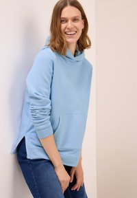 Giovane donna che sorride, indossando una felpa leggera blu e jeans blu scuro, appoggiata leggermente contro un muro bianco e beige.