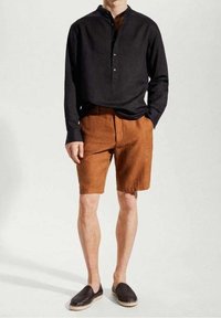 Hombre vestido con una camiseta de manga larga negra, pantalones cortos de color óxido y zapatillas negras sin cordones, de pie con una mano en el bolsillo.