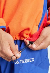 Flis jakna s dizajnom gradijenta u narančastoj i crvenoj boji; s plavim naglascima i podesivom crnom vezicom na rubu; logo na plavim šortsevima.