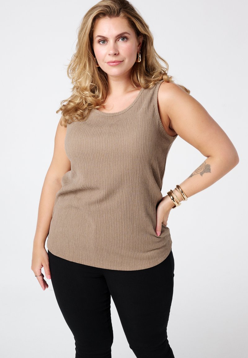Beige geribde tanktop met een ronde halslijn en mouwloos ontwerp, gecombineerd met zwarte skinny jeans. Gouden armbanden accentueren de pols.