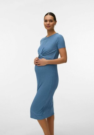 Zwangere vrouw in een blauw geribd gebreide jurk met korte mouwen, staand in profiel, zachtjes haar babybuik vasthoudend en glimlachend.