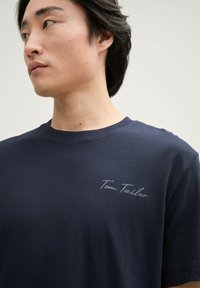 Tricou din bumbac bleu marin, cu guler rotund, mâneci scurte și un logo subtil "Tom Tailor" în gri deschis pe pieptul stâng.