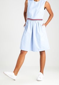 Helles blaues ärmelloses Kleid mit rundem Ausschnitt, Falten an der Taille und einem mit Rot akzentuierten Taillenbund. Die Modells steht in weißen Sneakers.