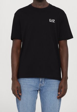 Mężczyzna w czarnej koszulce z logo EA7 Emporio Armani i jasnoniebieskich dżinsach, stojący na jednolitym tle.