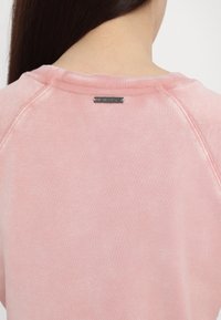 Hanorac roz deschis cu guler rotund și mâneci raglan. Etichetă de logo argintie notabilă la gulerul din spate. Material moale, cu textură.