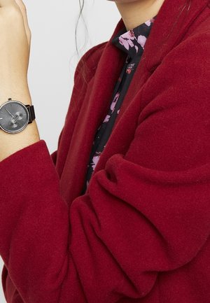 Femme portant un manteau rouge et un haut fleuri, montrant une montre-bracelet noire à cadran rond et bracelet en maille à son poignet gauche.