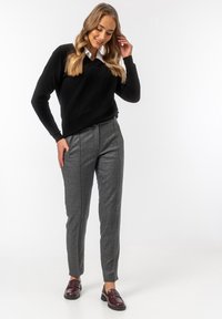 Czarny sweter z dekoltem w serek, szare dopasowane spodnie o teksturowanym wzorze oraz burgundowe mokasyny. Modelka stoi z jedną ręką na talii.