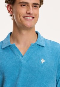 Gaiši zils frotē materiāla polo krekls ar V-veida kakla izgriezumu un maigu tekstūru, apdrukāts ar mazu balto izšūto logotipu krūtīs.