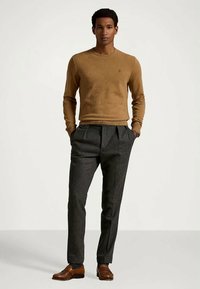 Άνδρας στέκεται με τα χέρια στα τσέπες, φορώντας ένα καφέ πουλόβερ, γκρίζες πτυχές παντελόνια και καφέ δερμάτινα παπούτσια σε απλό φόντο.