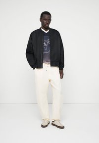 Veste bomber noire, pantalon oversize blanc et t-shirt graphique bleu marine. Les chaussures sont légères avec des accents texturés. Design simple et moderne.