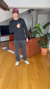 Svart langermet skjorte, grå jeans og hvite sko. Vist i et rom med tregulv, en plante og en oransje sofa i bakgrunnen.