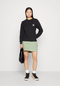 Schwarzes Langarmhemd mit einer Brusttasche, grüner Rock und schwarze Sneaker. Modell hält eine schwarze Tasche und steht vor einem schlichten Hintergrund.