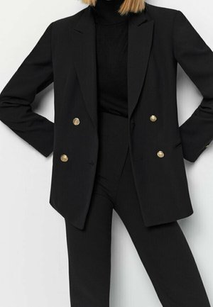 Femme portant un blazer noir croisé à boutons dorés sur un col roulé noir et un pantalon noir, debout les mains sur les hanches.