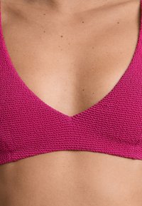 Fuchsiový texturovaný bralet s hlubokým výstřihem do V. Tkanina vypadá jako tkaná, má hladký povrch a čisté okraje. Žádné viditelné kovové prvky.