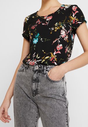 Kobieta w czarnej, krótkim rękawie, kwiatowej bluzce włożonej do szarych, wysoki stan, jeansów acid wash, z jedną ręką w kieszeni.