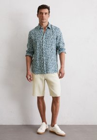 Chemise boutonnée bleu clair avec un motif floral, short crème et baskets blanches. La chemise a des manches retroussées et une coupe décontractée.