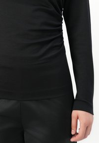 Svart langermet topp med rynkede sidelagsdetaljer, båret med sorte bukser, som viser høyre side av en persons torso og hånd.