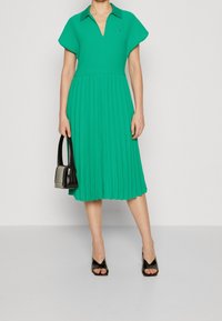 Vihreä laskostettu mekko, jossa on kauluspanta. Lyhyet hihat ja V-kaula-aukko. Asusteena musta käsilaukku ja korkokenkiä.