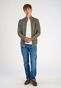 Green textured cardigan with high collar, worn over a white shirt, paired with blue jeans and brown boat shoes. Casual fit and comfortable design.