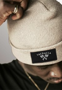 Beige knitted beanie with a textured pattern and black label featuring the word "valhalla" and crossed axes. Close-up view.