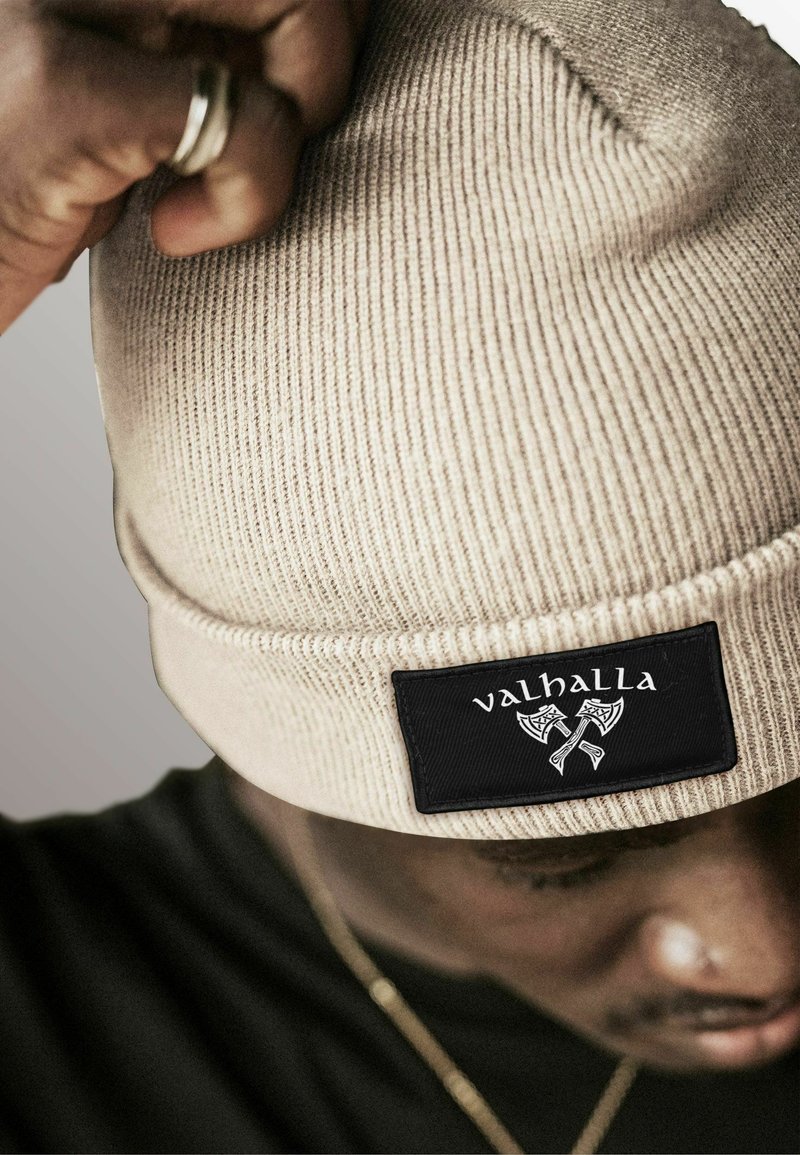 Beige knitted beanie with a textured pattern and black label featuring the word "valhalla" and crossed axes. Close-up view.