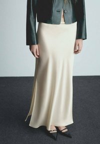 Krémová saténová maxi sukně se bočním rozparkem, zkombinovaná s černou koženou krátkou bundou a černými lodičkami s špičatou špičkou. Hladká textura.