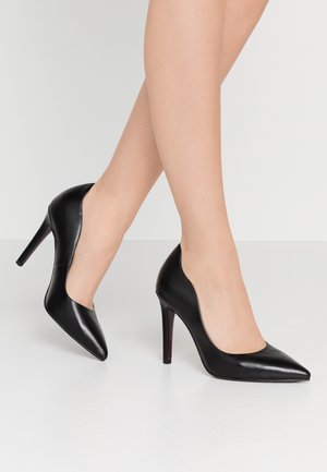 Jambes portant des escarpins noirs pointus à talons aiguilles fins sur un fond clair uni.