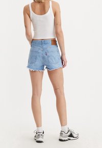 Pantalones cortos de mezclilla azul claro con dobladillo desgastado, dos bolsillos traseros y una etiqueta de cuero marrón. Combinados con una camiseta sin mangas blanca y zapatillas deportivas.