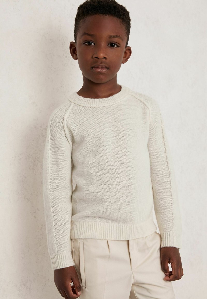 Junge mit kurzen lockigen Haaren, der einen cremefarbigen Strickpullover und beigen Hosen trägt, steht vor einer hellen strukturierten Wand.