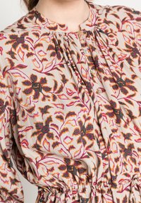 Blommig blus i ljus beige med orange, rosa och svarta mönster. Har en plisserad halsringning och knäppning. Elastisk midjedetalj.