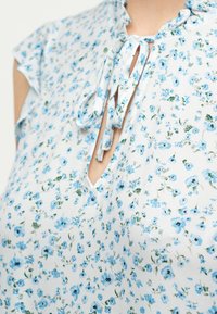 Κοντινή λήψη από ελαφρύ ύφασμα με μικρό μπλε λουλουδάτο σχέδιο και λαιμόκοψη με άνοιγμα που δένει με δύο υφασμάτινα κορδόνια.